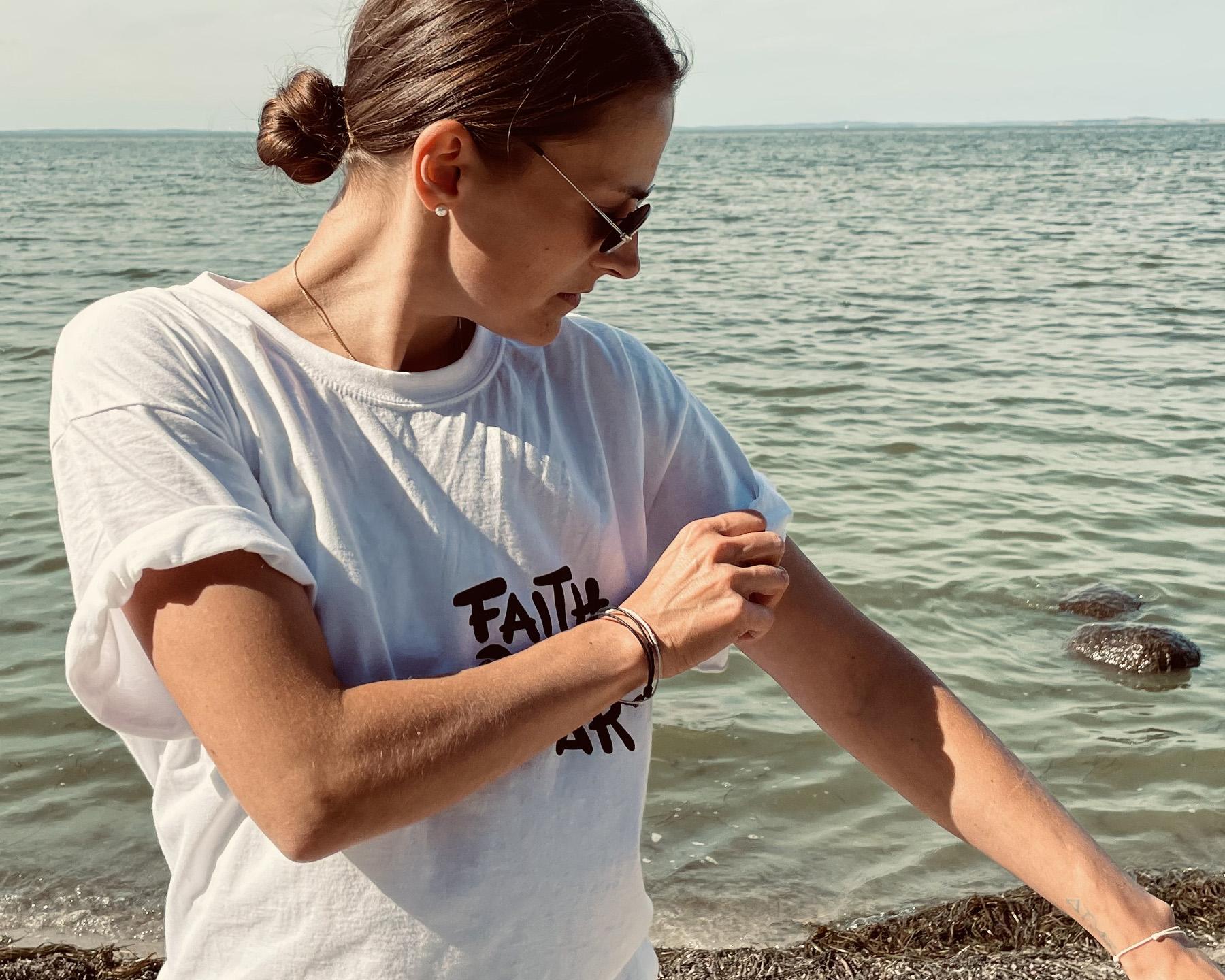 Frau mit weißem Faith Over Fear T-Shirt und Sonnenbrille vor dem Meer. Dein Mindfulness Store.